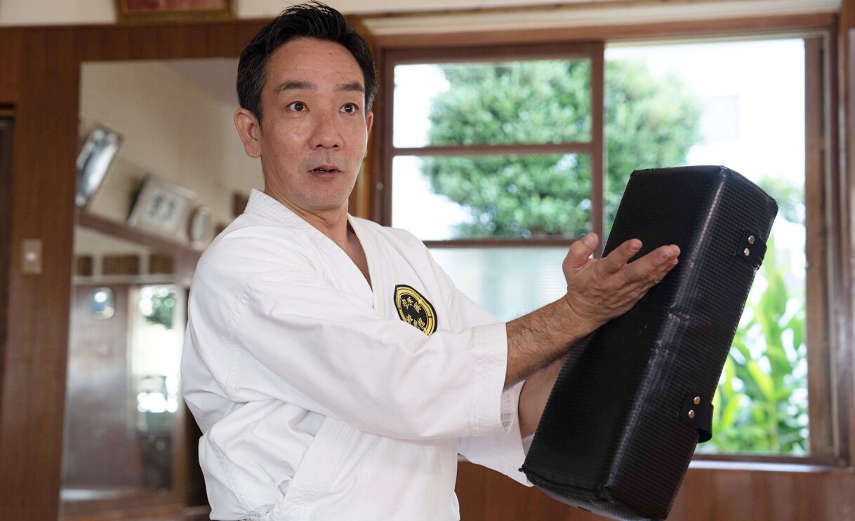 An image of a man teaching karate in Shorin-ryu Seibukan, one of Okinawa's traditional karate schools. He is holding a back block in one hand and pointing to it with the other. 