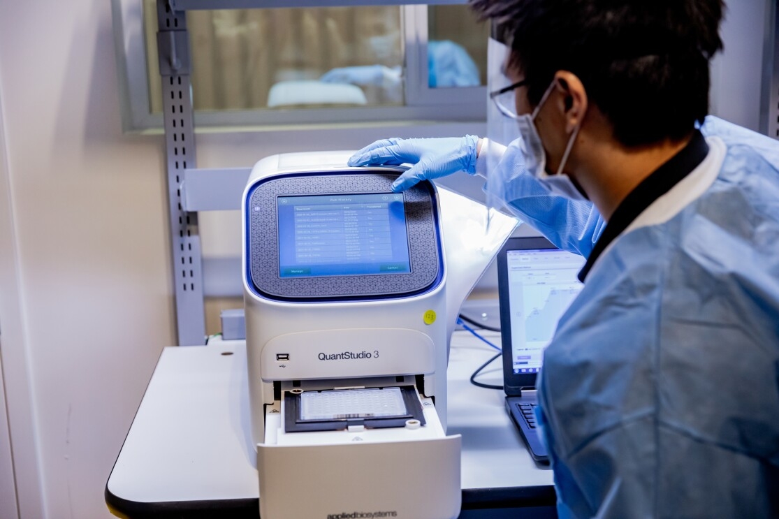 A man wearing protective gear, including a face shield, mask, gloves, and protective clothing touches the screen on a piece of laboratory equipment.