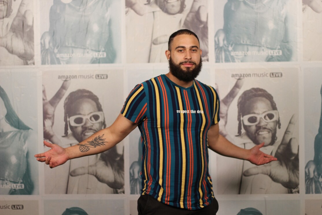 A headshot image of Julian Ortiz in front of a backdrop of Amazon Music Live show posters featuring Anitta and 2 Chainz.