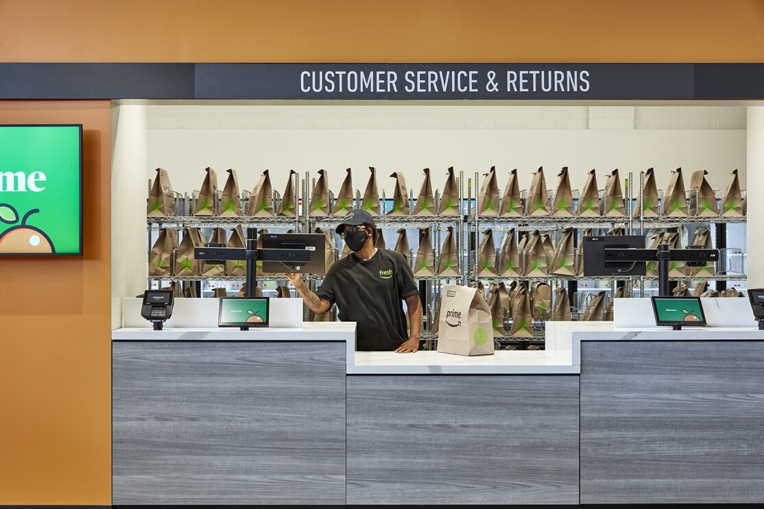 An image of an employee at an Amazon Fresh store working behind the counter of a customer service desk. There are bags with grocery deliveries behind her.