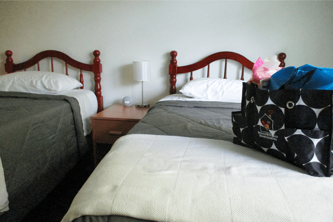 A guest suite at the Ronald McDonald House in Washington, D.C. has two twin beds with a nightstand in between them. On the night stand is a lamp and an Amazon Echo. There is an overnight bag with the Ronald McDonald House Charities logo on it.