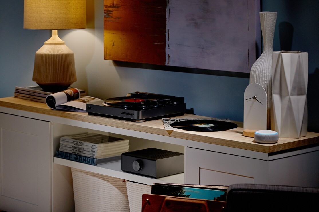 An Echo Link Amp in a living room setting. Surround it are magazines, records, a record player, lamp, decorations and Echo Dot device.