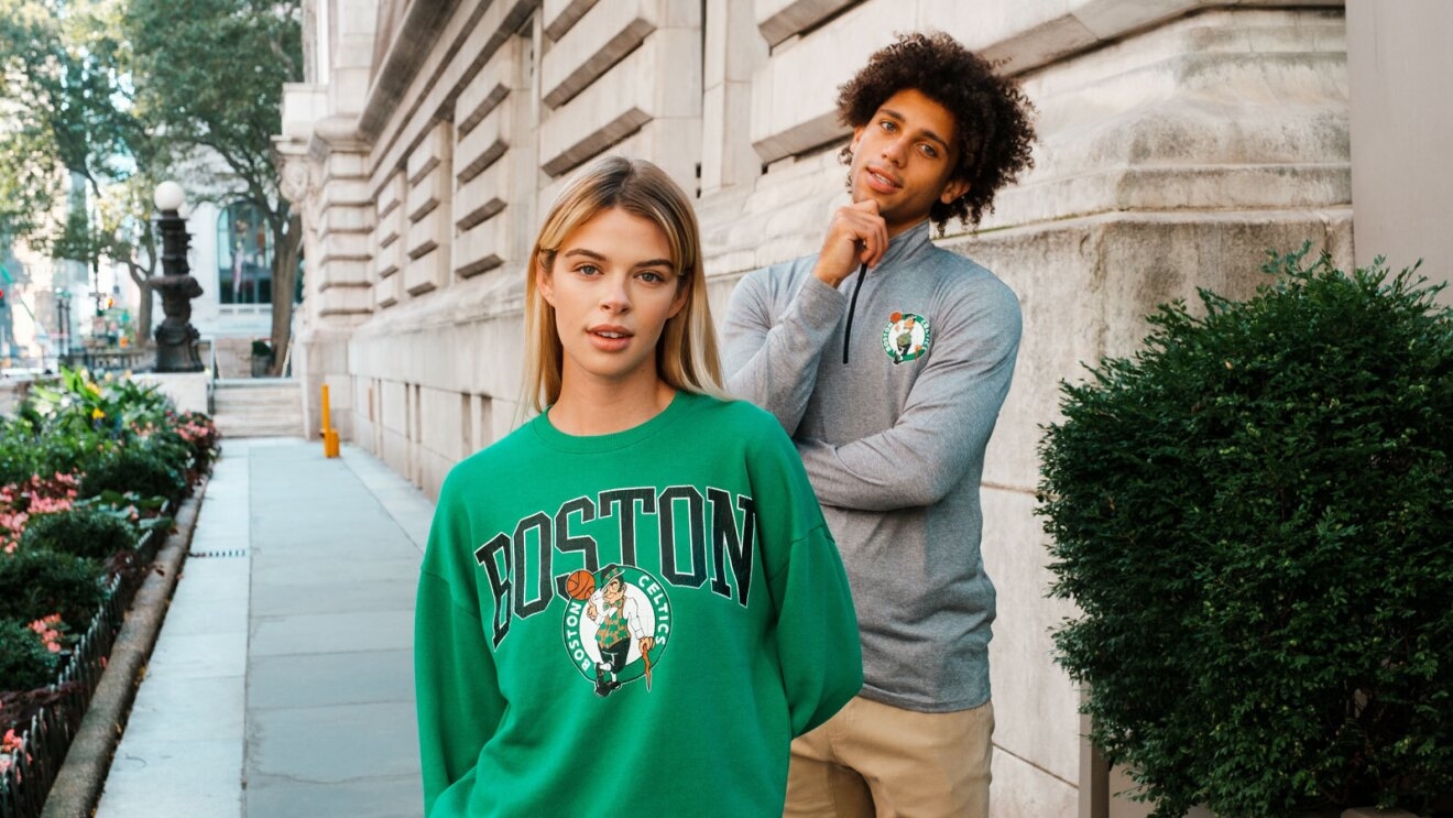 An image of a man and a woman. The woman is wearing a green pullover sweater with the word "Boston" written on the front and the Celtics logo. The man is wearing a gray, quarter zip long-sleeve shirt with the Celtics logo on the left chest.