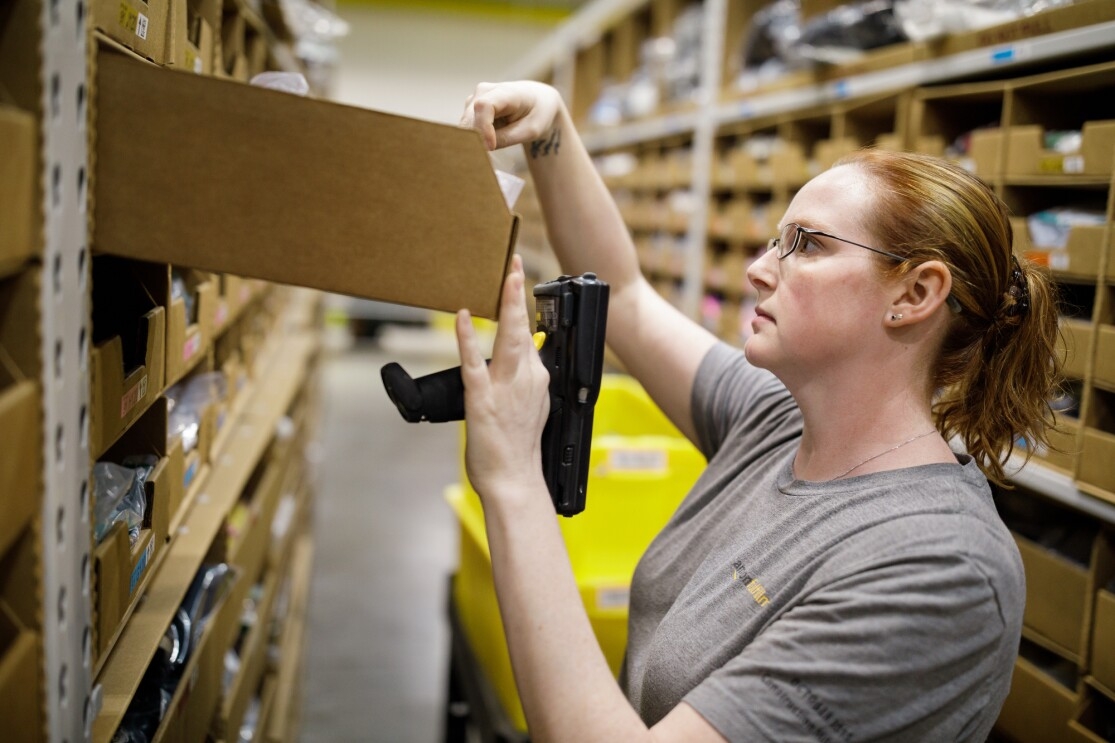 A woman with red hair holds a scanner and looks inside an open cardboard bin. 