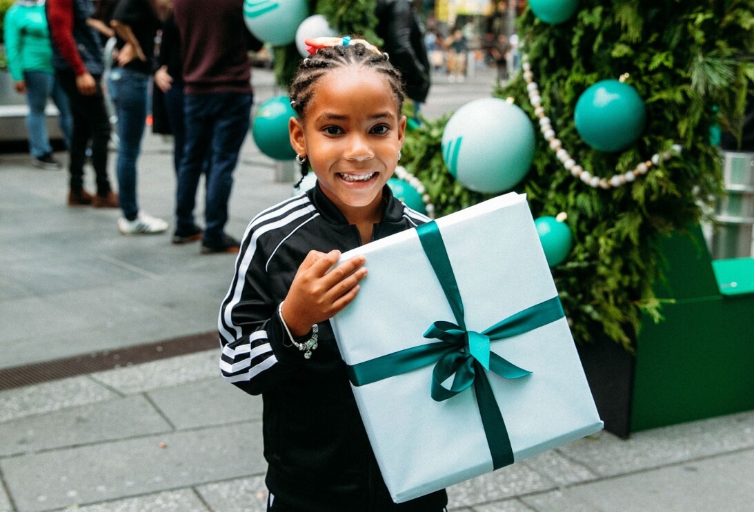 An image of a young girl smiling for a photo while holding a wrapped present. There is a restive wreath in the background of the photo. 