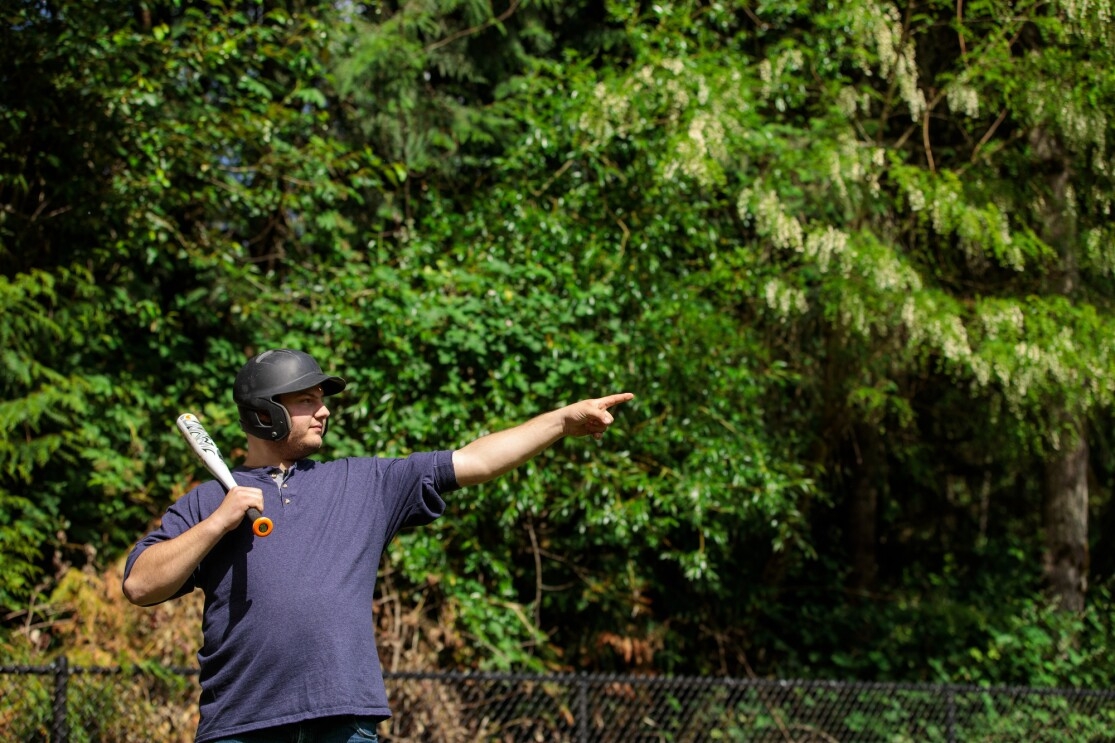 A man balances a baseball bat on his shoulder. He wears a batting helmet. With the hand that's not holding the bat, he points up and away toward the spot he plans to hit the ball.