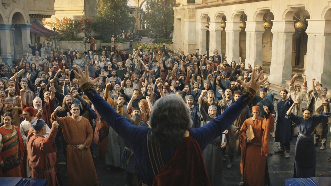 A crowd of people joyously shout at the leader who's facing them with his arms up in the air. Everyone is standing in a plaza of marble buildings.