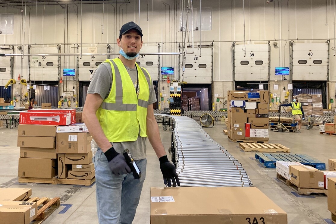 A man wearing a safety vest and a protective mask stands next to a stack of boxes holding a barcode scanner.