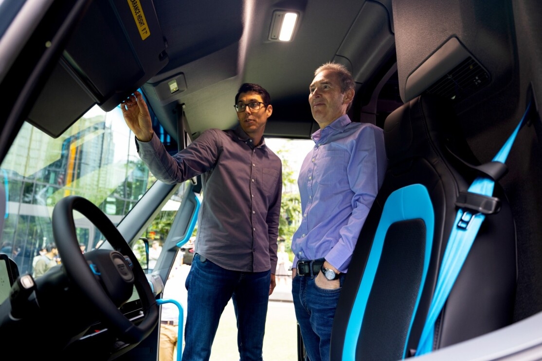 Andy Jassy and RJ Scaringe stand in a Rivian Amazon delivery vehicle that is parked outside Amazon headquarters. Scaringe points to a part of the innovative design on the driver side.