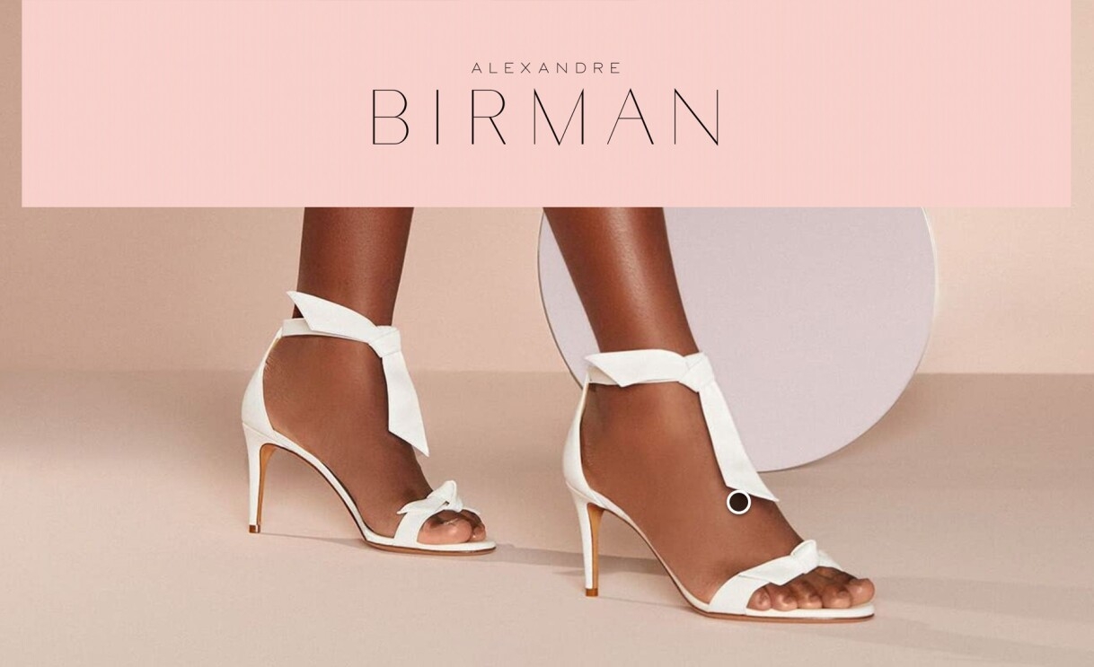 An image that shows white heels worn by a model and text that reads "Alexandre Birman" at the top. 