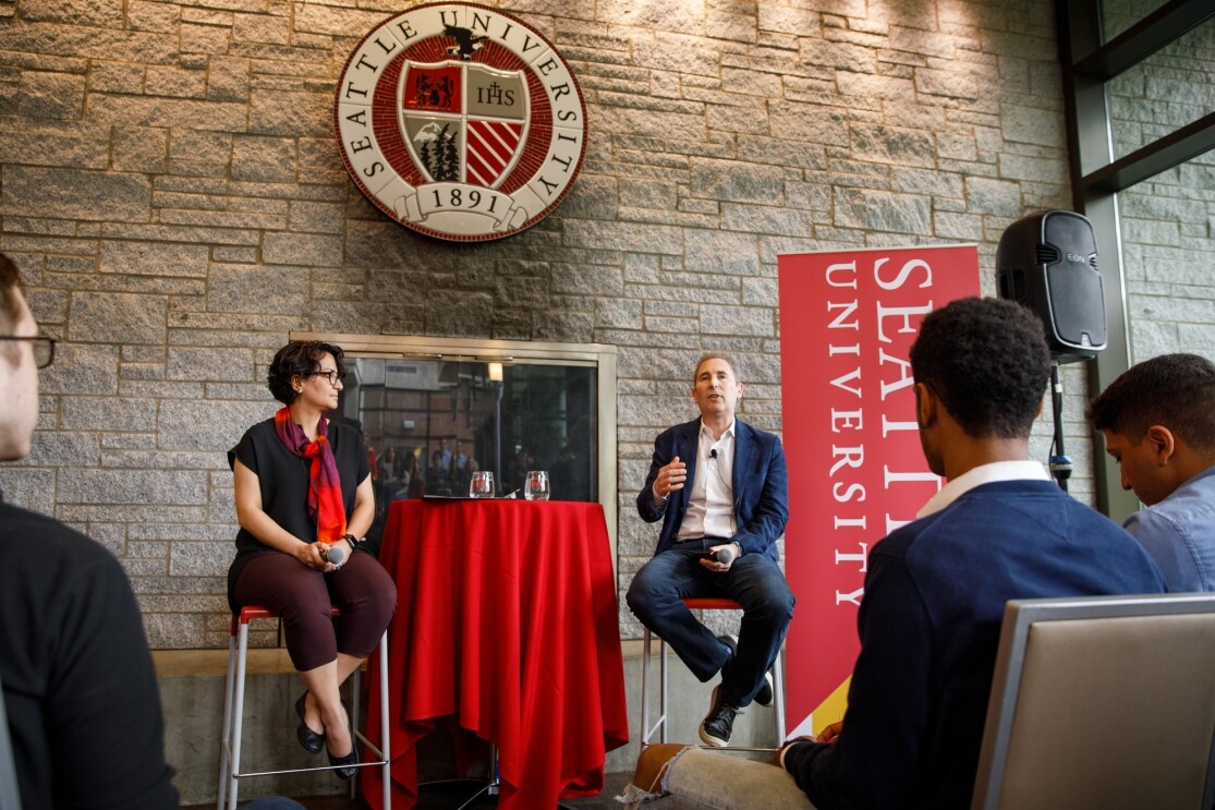 A woman, Roshanak Roshandel sits on a raised chair, next to a man, Andy Jassy, as they lead a fireside chat at Seattle University.