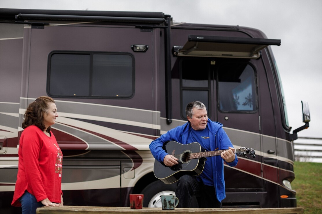 Flanked by a motorhome, a woman watches a man play guitar.
