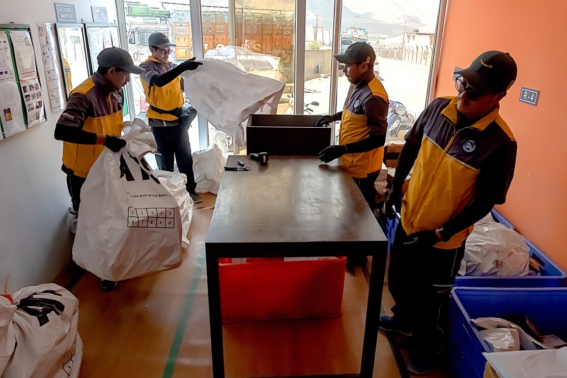 Four men in uniforms with Amazon logos stand around a table. One man carries a large plastic bag. Another man shakes an empty plastic bag in preparation for leading it. Bins and packages are on the floor at the right side of the image.