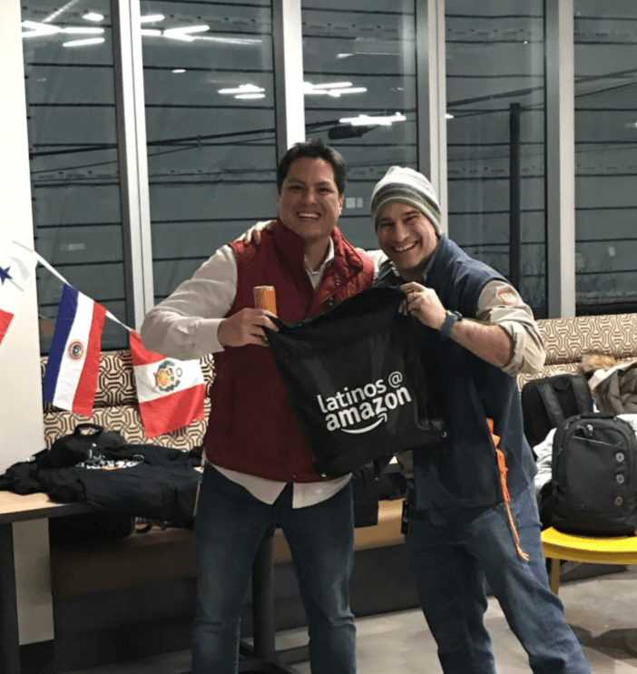 Two Amazon employees smile while holding up a backpack with the Latinos@ Amazon logo on it.