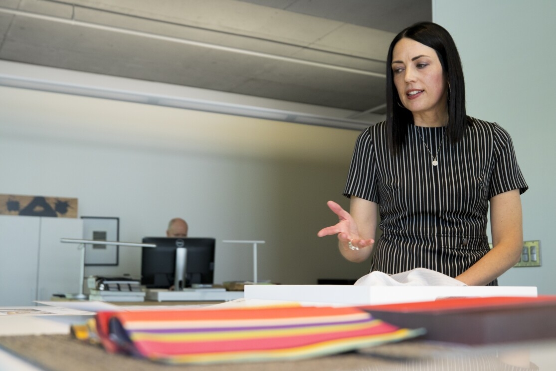 A woman in a grey-striped shirt stands over architectural designs. Her colleague is in the background at a computer.