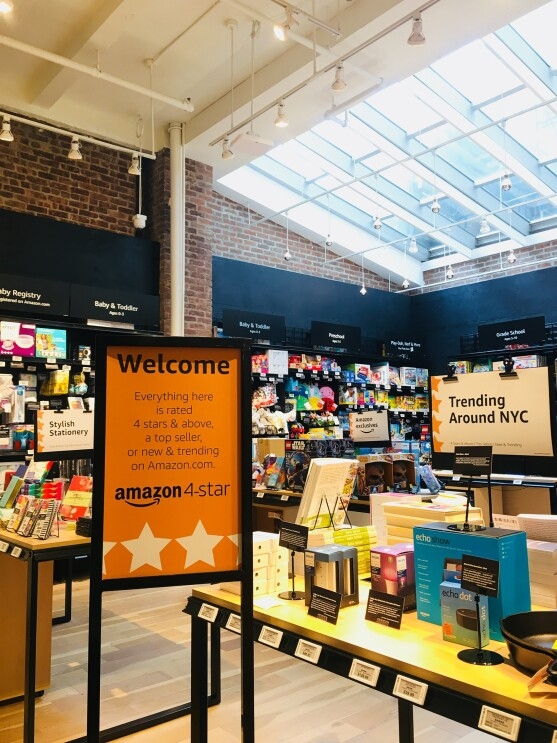 View inside Amazon 4-star. In the foreground, a sign that says "Welcome Everyting here is rated 4 stars & above, a top seller, or new & trending on Amazon.com. Next to the sign is a display table with products on it, and a "Trending Around NYC" sign. Some items on the table include Echo Show, Echo Dot, books and more. In the background, baby registry, baby and toddler products, grade school items, and more. 