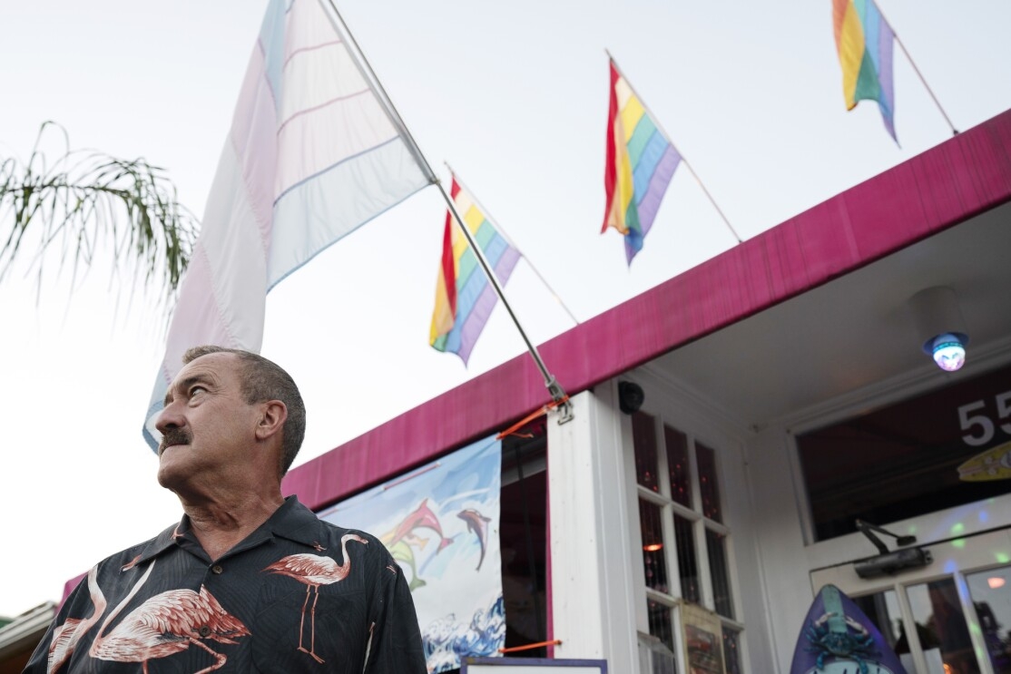 A man stands in front of a building flying gay pride and transgender pride flags.