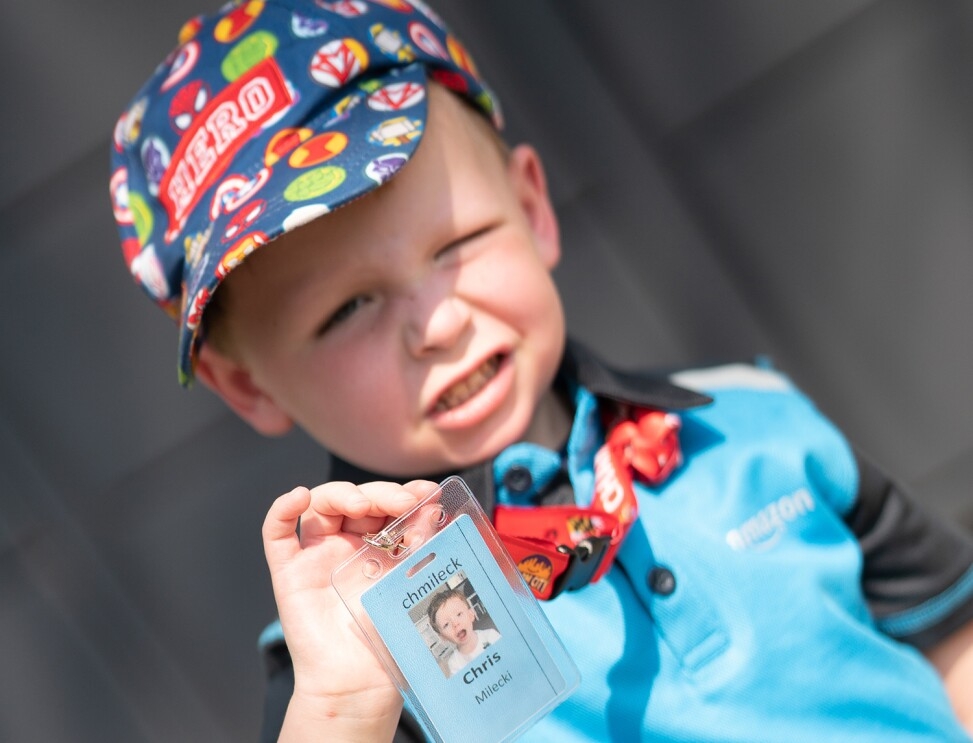 An image of a boy with a colorful hat on that says "hero" smiling for a photo while showing an Amazon badge with his name on it and wearing an Amazon shirt. 