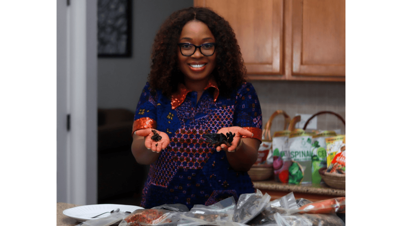 Toyin Kolawole, CEO, Iya Foods holds up some of her product while she smiles for the camera. In front of her and behind her are bags of her product. She wears a blue patterned dress with an orange collar and sleeves. She has dark curly hair to her shoulders and wears black-framed eyeglasses.