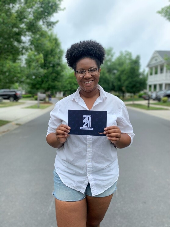 An image of a woman smiling for a photo on a street in her neighborhood while holding a paper invitation to Amazon's 2021 internship class.