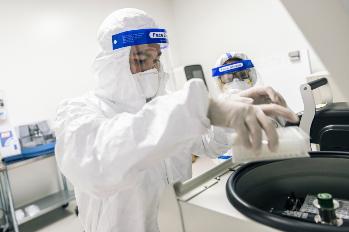 Two people in PPE. The person in the foreground places a tray inside a piece lab equipment.