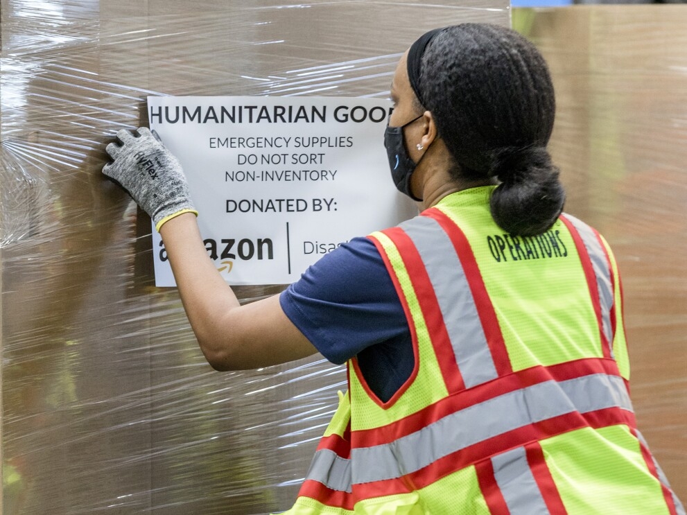 A woman wearing a mask navigates a pallet of humanitarian goods in an Amazon fulfillment center
