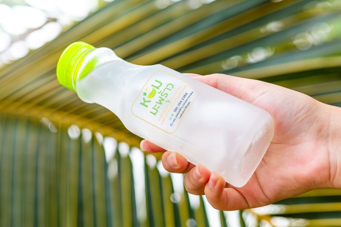 An image of a hand holding a clear botte with a green cap and a green label on it. The contents are coconut water. There is a palm leaf in the background of the image.