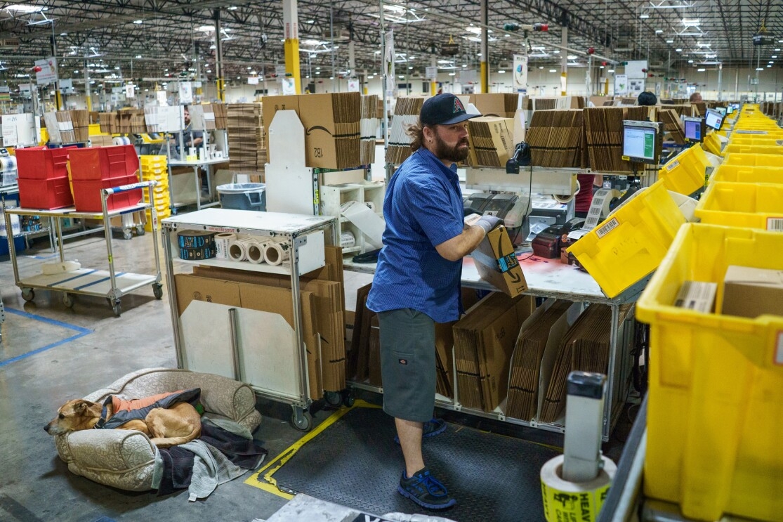 A man in a baseball cap handles a cardboard box marked with the Amazon logo. A dog rests in a small dog bed placed a few feet behind the man. In the background, there is a large warehouse space.