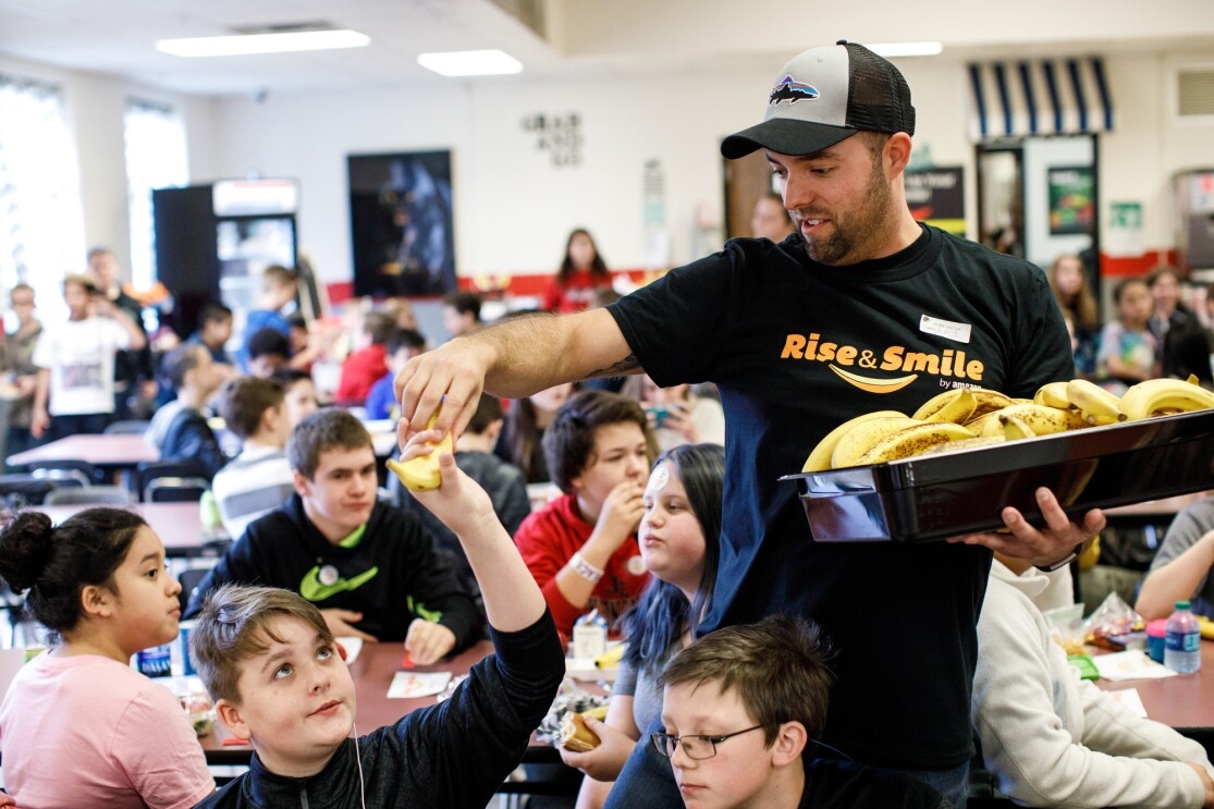 A man holds a tray of bananas and wears a Rise & Smile tee as he hands a banana to a student at a school