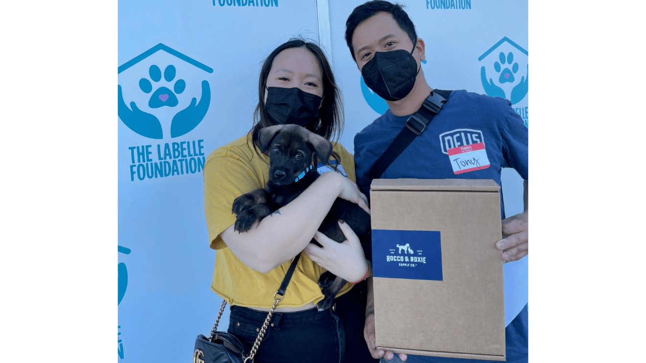 Two people pose for a picture with their new puppy. One of them is holding a Rocco & Roxie box while the other holds the black and brown puppy close.