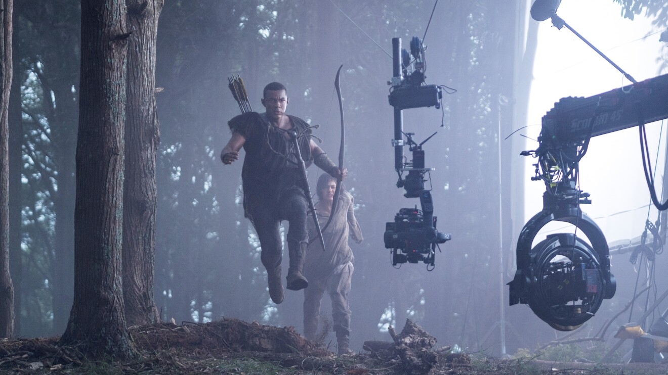 Behind-the-scenes view of two characters running through a forest, one of which is jumping mid-air while holding a bow and arrow.