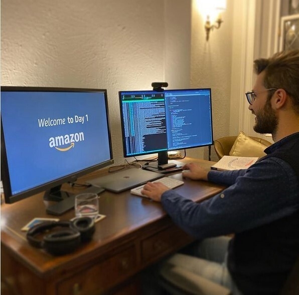 An image of a man smiling while working at his computer. One monitor has code on the screen and the other has the Amazon logo.