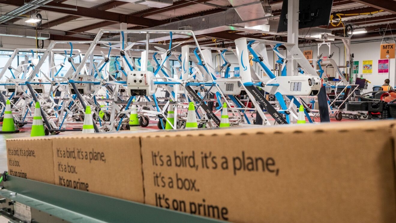 A row of boxes inside the Amazon drone facility. The text on the boxes says "it's a bird. it's a plane. it's a box. it's on Prime." There are several rows of delivery drones in the background.