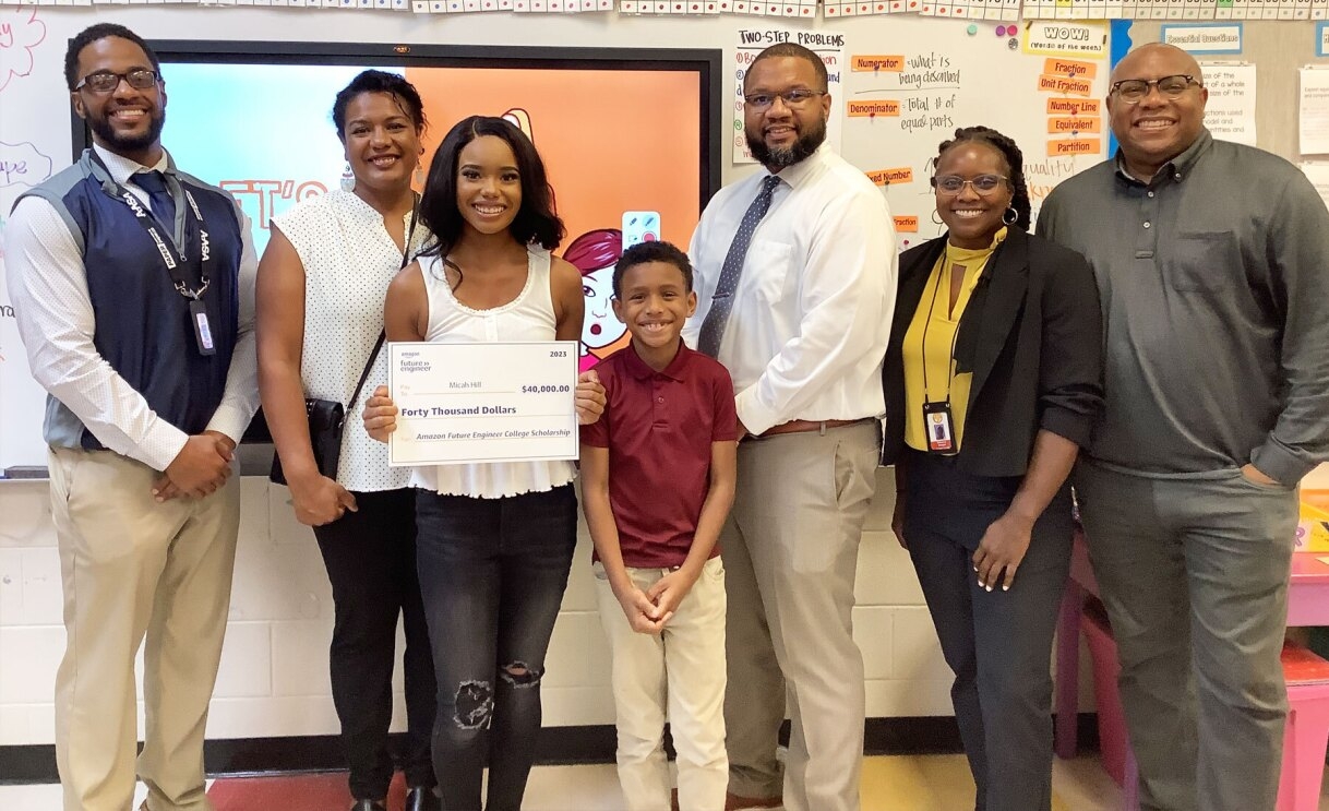 An image of Micah Hill and her family smiling to the camera and holding out a check for the 2023 Amazon Future Engineer Scholarship.