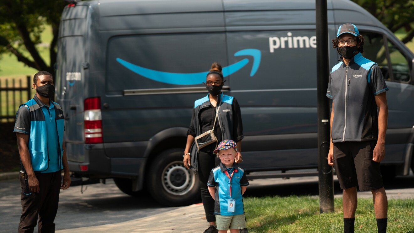 An image of a boy standing in front of an Amazon van while wearing an Amazon employee vest. There are two employees standing for the photo with him.