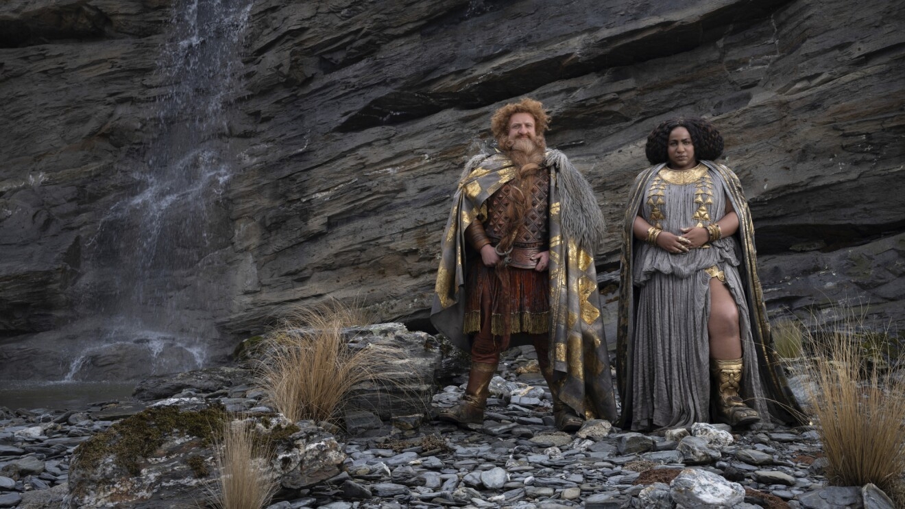 A man and a woman dressed in garments of grey cloth and gold pose for the camera in a rock-y terrain.