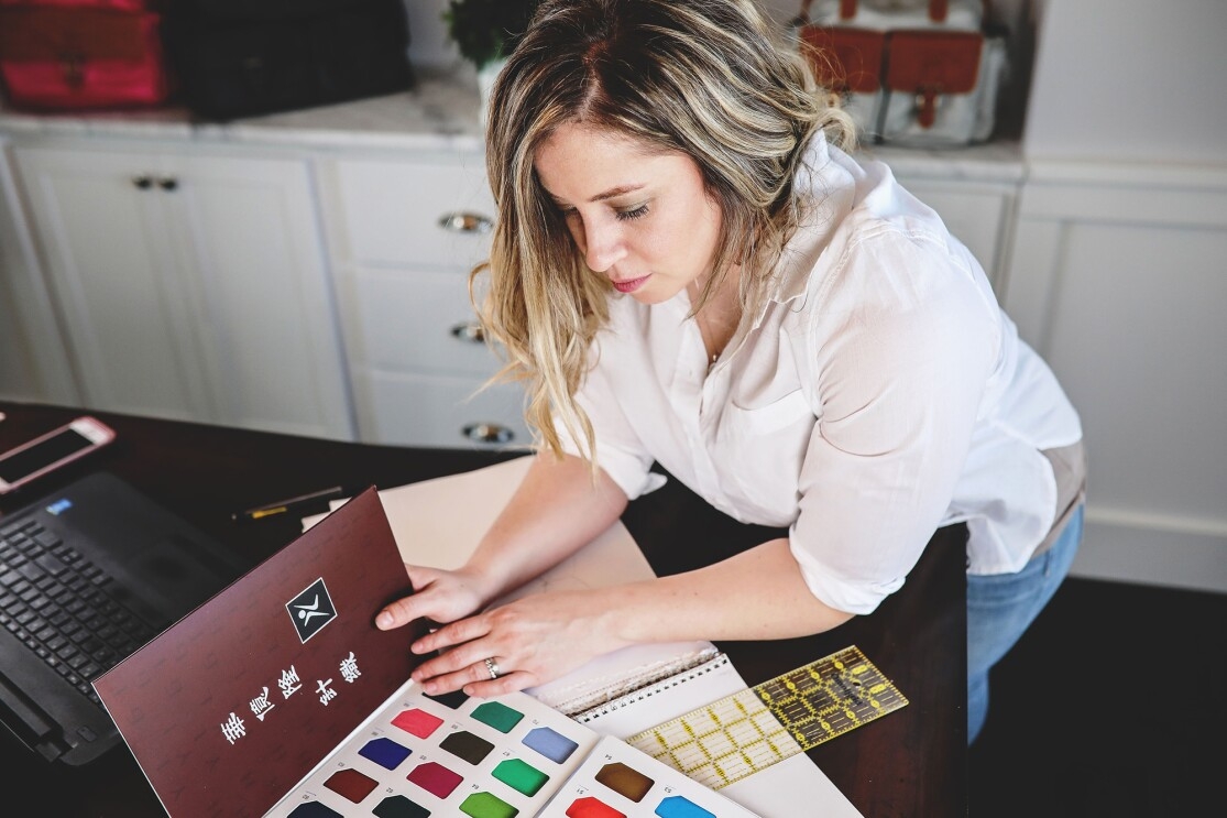 A woman wearing a white button down and blue jeans holds a fabric swatch book, examining the contents of the book. She leans over a desktop, which also holds her computer, notebook, and other office supplies.