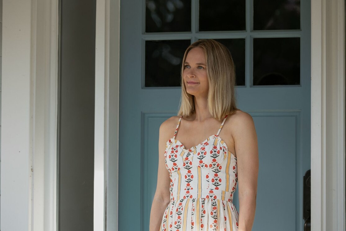 Susannah from "The Summer I Turned Pretty" stands on her porch in a white sundress with colorful patterns looking at something off camera. 