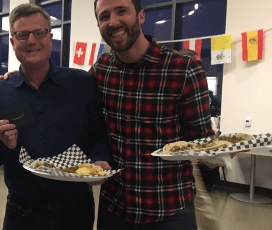 Two men smile for the camera while holding plates with tacos on them. 