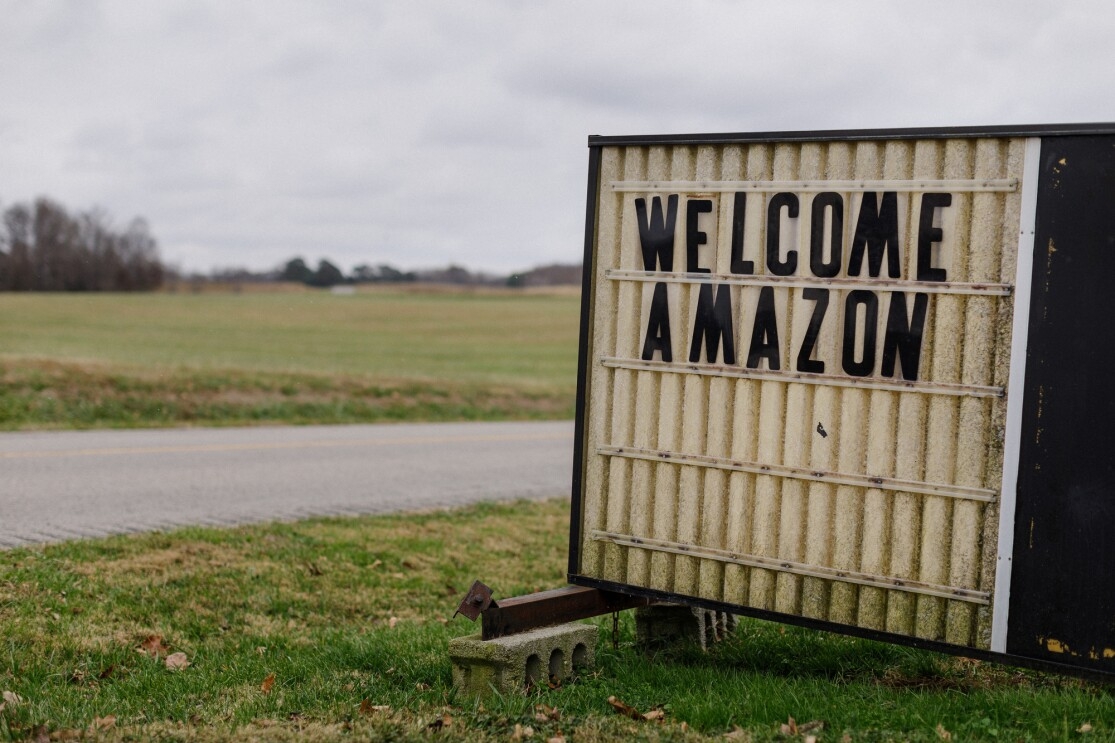 The message 'WELCOME AMAZON' is displayed on a sign balanced atop cinder blocks sitting on a lawn. In the background of the image, there's a road and a grassy field.