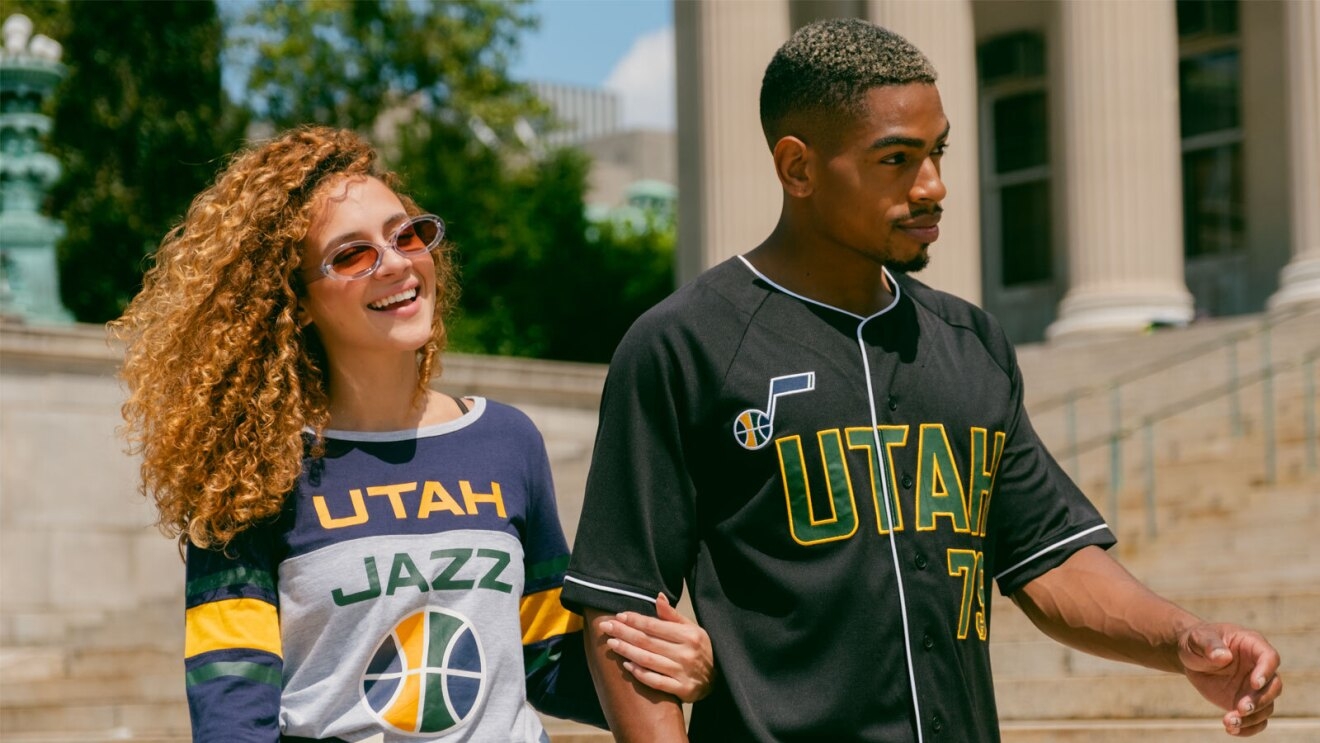 An image of a man and a woman walking arm-in-arm in front of a building with a large, cement staircase. The woman is smiling wearing a grey, navy blue, yellow, and green long-sleeved shirt with the words "Utah Jazz" on it and a basketball below the wording. The man is wearing a black baseball jersey with the word "Utah" on the front in green lettering with a yellow outline, and the Jazz logo (a music note in navy blue) in the top corner.