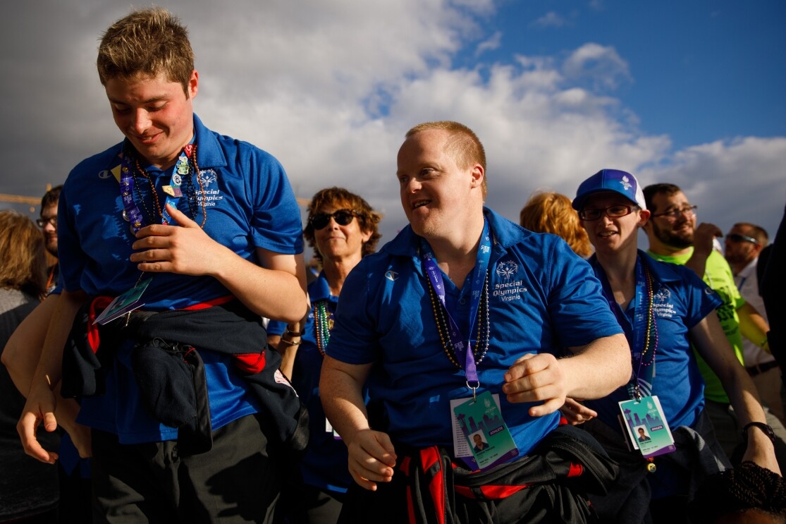 Special Olympics athletes and attendees from Virginia appear to lead a group of others. Two smiling males are in the foreground, seeming to be moving forward. In the middle ground, two other individuals wearing "Special Olympics Virginia" shirts are facing the same direction. In the background, more attendees are seen.