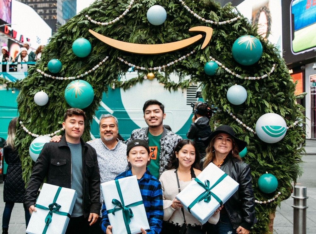 An image of a family smiling for a photo in front of a wreath. The kids are holding presents with white wrapping and green bows.