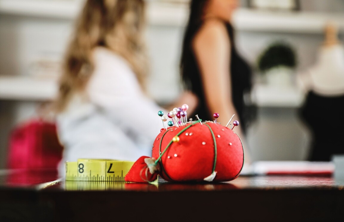 A pin cushion with sewing needles embedded in it sits in the foreground of the image, next to a measuring tape used for sewing. In the background, a blond woman pins the back of a black dress being worn by a customer.