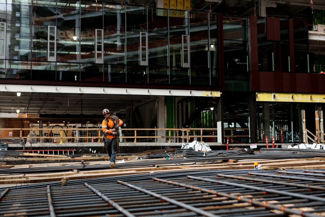 A man in a construction helmet and orange safety jacket is photographed at a construction site on Amazon's campus in Seattle, WA. He is walking across an unfinished floor covered in rebar.