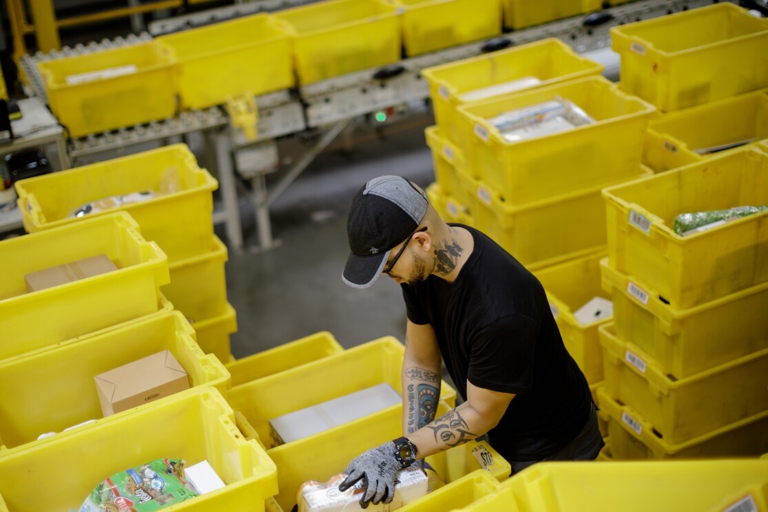 A man stands among yellow plastic tote boxes and handles merchandise. He's wearing a baseball cap, glasses and work gloves, and has tattoos on each arm and his neck.