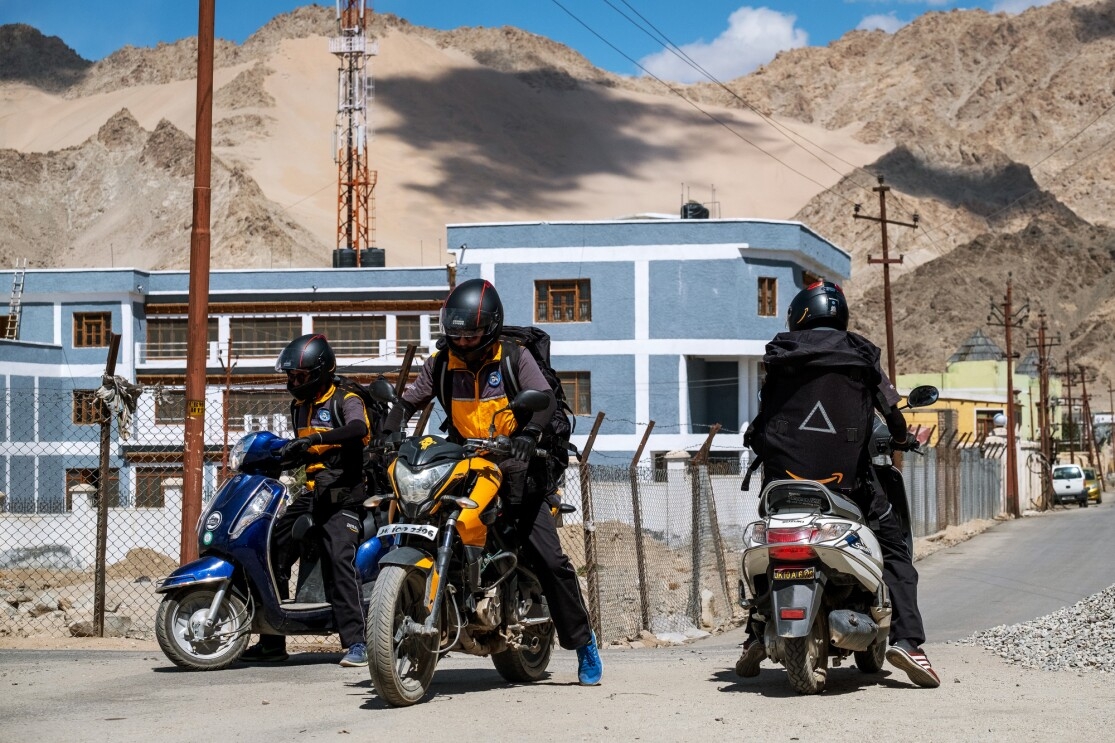 On a dirt road, one man sits on a motorcycle, and two men sit on motor scooters. All three men wear helmets, backpacks, and uniforms. They appear to be preparing to ride away.