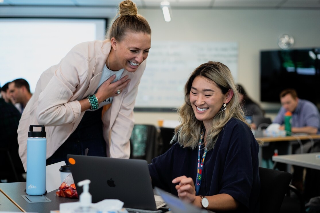 Two women, one sitting and one standing, share a smile as they both look at a computer in a classroom setting.