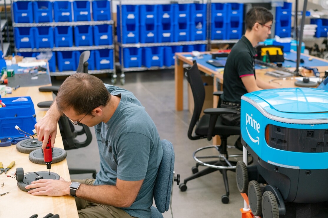 A man uses a tool as he works on a wheel for Scout. Behind him sits a Scout robot, and another man is seen working as well.