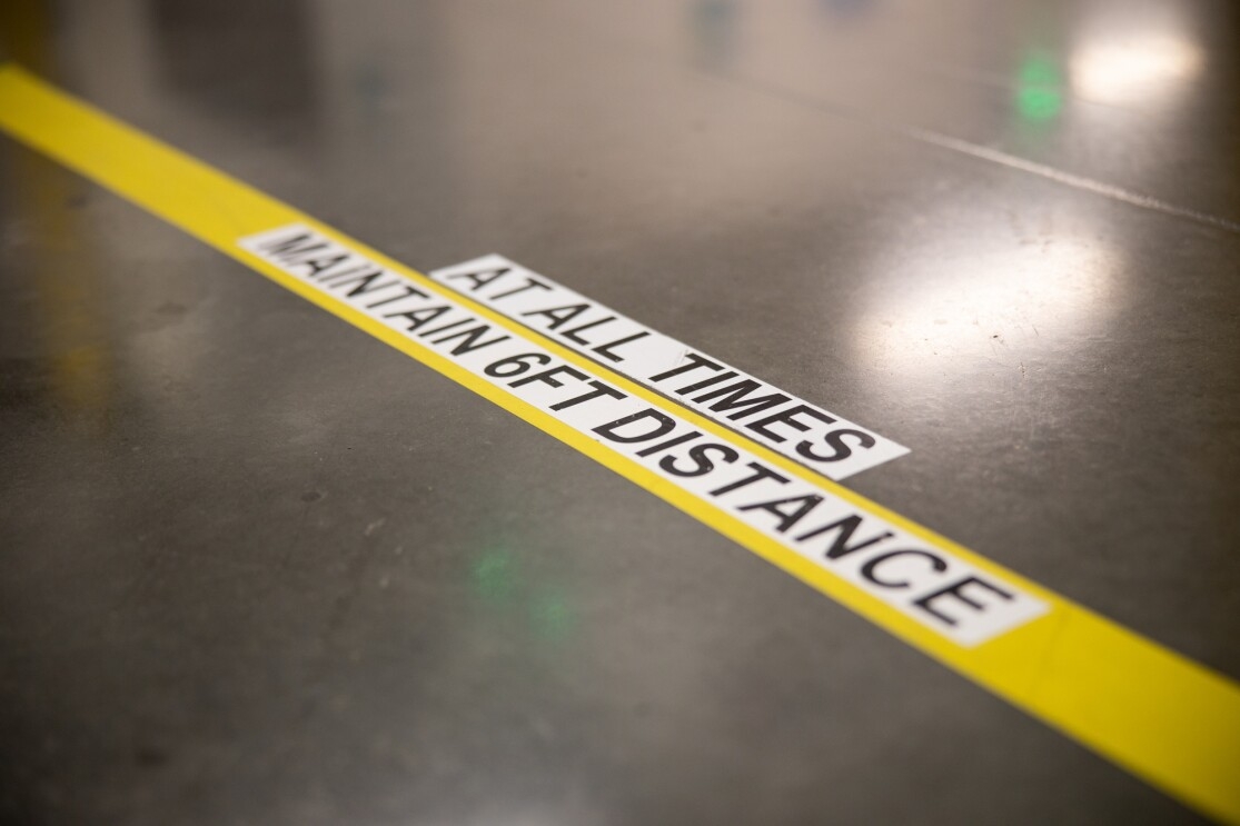 Markings on a concrete floor in a workplace say "MAINTAIN 6FT DISTANCE AT ALL TIMES."